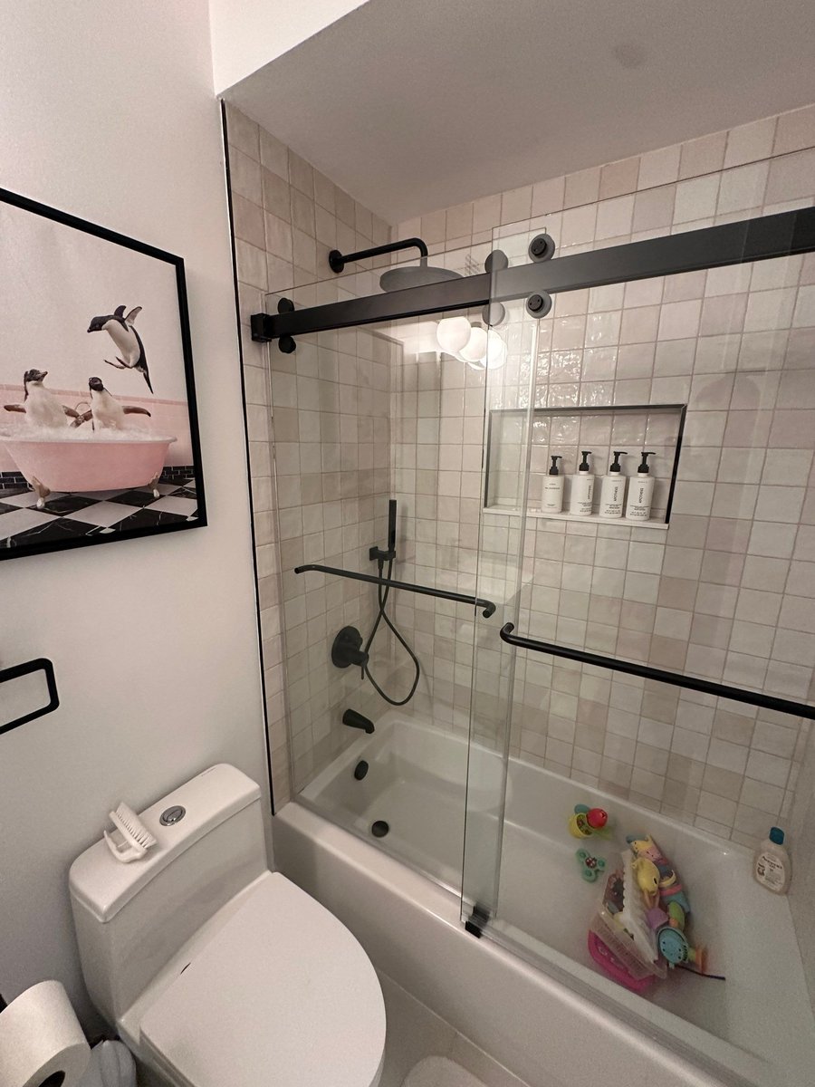 apartment renovation bathroom with black framed sliding glass shower over bathtub white square tile walls and toilet in manhattan valley ny by modo renovations